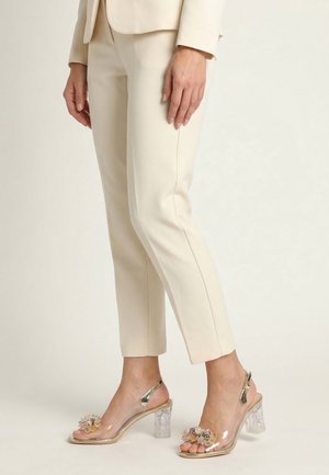 Vrouw draagt crème-kleurige op maat gemaakte broek en transparante sandalen met blokhakken en bloemversieringen op de tenen.