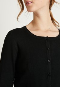 Cardigan nero con scollatura rotonda, realizzato in tessuto liscio, con chiusura frontale a bottoni e texture a coste lungo i bordi.