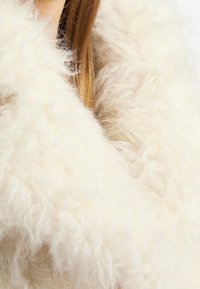 Puchaty, biały szalik z gęstym, teksturowanym materiałem przypominającym futro. Otaczające włókna tworzą efektowny, objętościowy wygląd. Miękki i ciepły w dotyku.