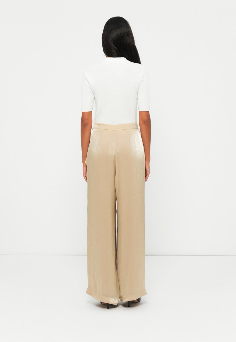 Beige, weite Hosen mit glänzender Textur, kombiniert mit einem figurbetonten weißen T-Shirt mit kurzen Ärmeln, von hinten vor einem weißen Hintergrund gezeigt.