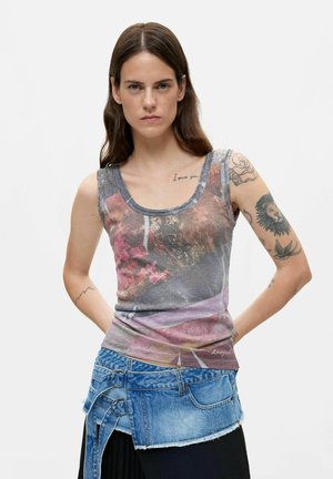 Donna con lunghi capelli castani che indossa una canotta floreale senza maniche e una gonna di jeans asimmetrica con anelli metallici, in piedi con le mani dietro la schiena.