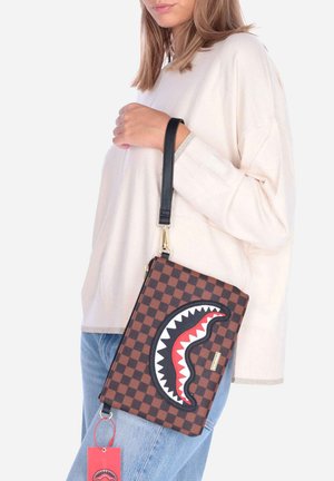 Mujer con suéter crema y vaqueros azules sostiene un bolso de mano marrón a cuadros con un diseño de boca de tiburón en negro, rojo y blanco y una correa negra.