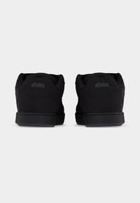 Ζευγάρι μαύρα αθλητικά παπούτσια Etnies χαμηλού προφίλ, εμφανιζόμενα από πίσω, με επενδυμένους γιακάδες και λαστιχένιες σόλες σε απλό φόντο.