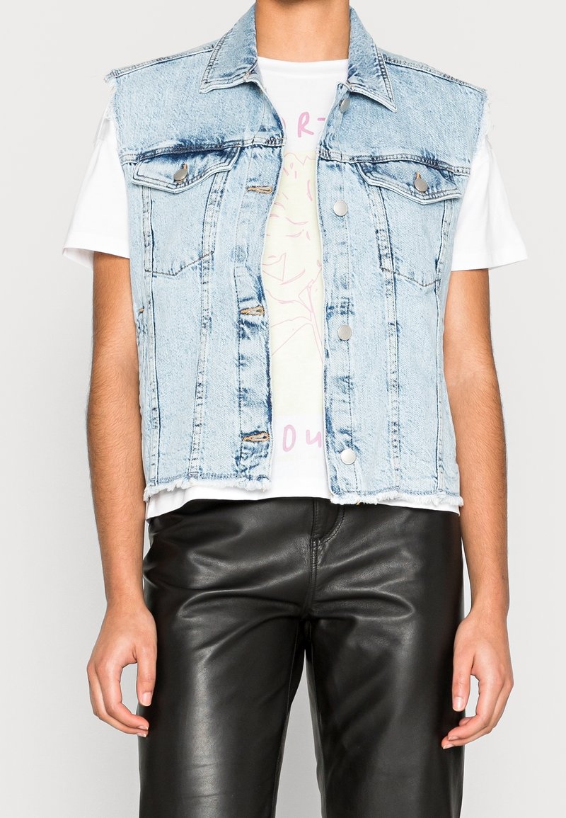 Jasnoniebieska kamizelka denimowa z surowym dołem, dwoma przednimi kieszeniami i metalowymi guzikami, noszona na białym T-shircie z pastelowym wzorem, w zestawie z czarnymi spodniami.