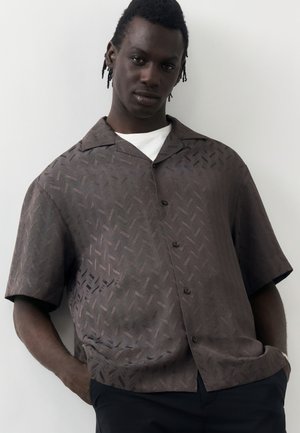 Ung mann med korte dreadlocks iført en brun mønstret kortermet skjorte over en hvit T-skjorte, hendene i lommene.