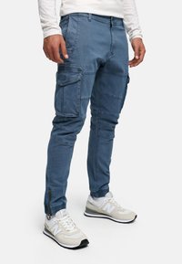 Blå cargo bukser med flere lommer, struktureret stof og smal pasform. Har lynlåse ved anklerne og en taljebånd med bæltestropper.