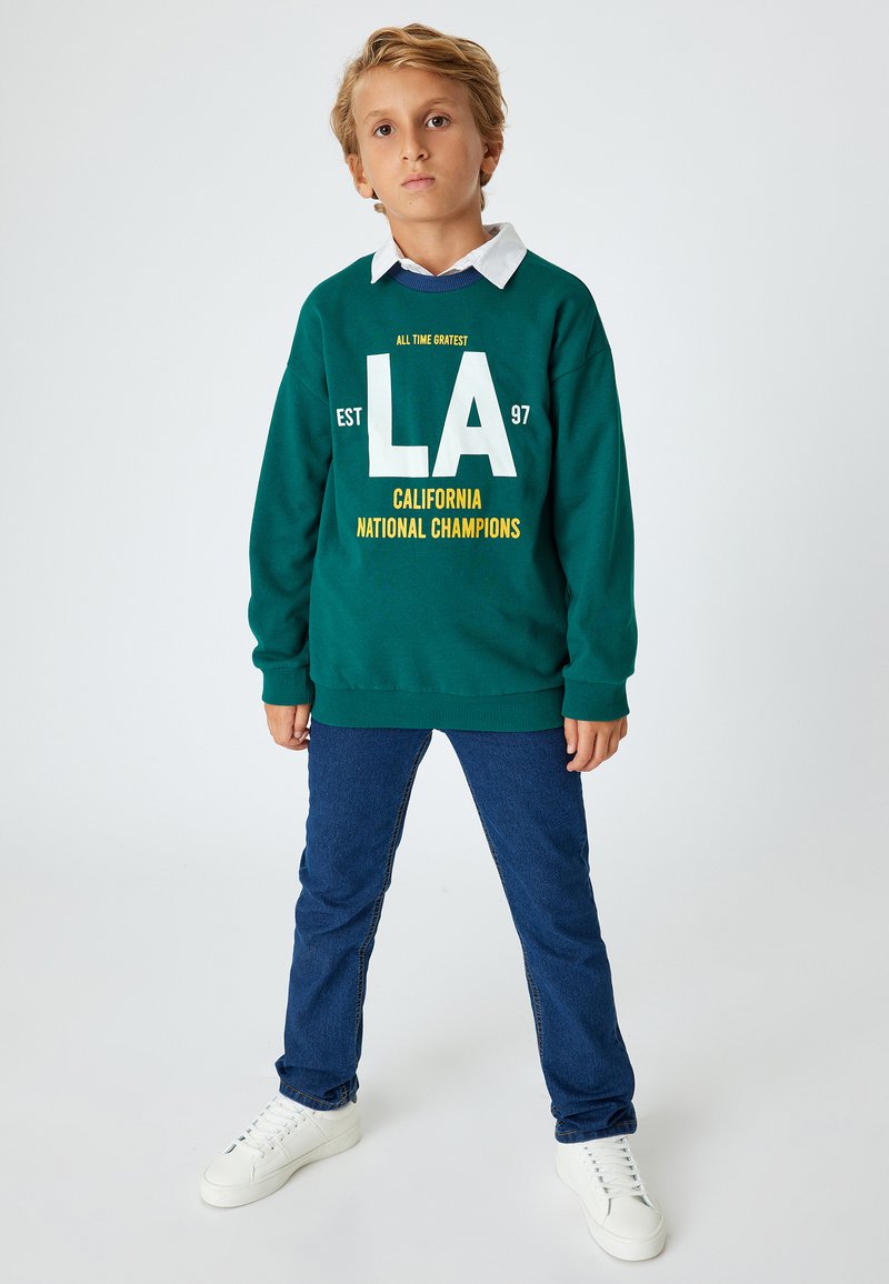 Sudadera verde con texto gráfico en blanco y amarillo, jeans azules y zapatillas blancas. Características notables incluyen un cuello y puños de manga acanalados.