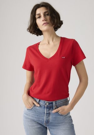 Kobieta o krótkich brązowych włosach, ubrana w czerwony t-shirt z dekoltem w serek i jasnoniebieskie dżinsy, stojąca z rękami w kieszeniach na jednolitym tle.