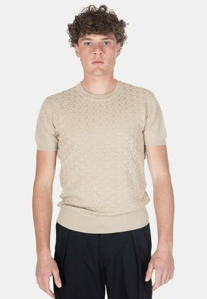 Giovane uomo con capelli ricci che indossa un maglione beige a maniche corte con trama e pantaloni neri, in piedi davanti a uno sfondo bianco uniforme.
