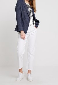 Blazer doppiopetto blu navy sopra una camicia a righe bianco e nere, abbinato a jeans bianchi cropped e sneakers bianche.