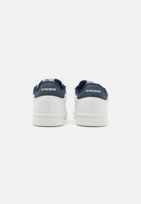 Baskets blanches avec une finition en cuir lisse, des accents bleu marine et un talon texturé. Présente le logo "le coq sportif" à l'arrière.