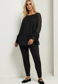 Top nero a maniche lunghe con polsini a volant e decorazioni di perline, abbinato a pantaloni neri attillati e scarpe nere flat. Tessuto liscio.