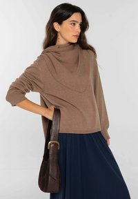 Suéter marrón de punto con escote drapeado, mangas largas y puños acanalados, combinado con un vestido azul marino y un bolso de cuero marrón.