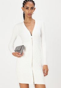 Robe blanche côtelée avec une fermeture éclair à l'avant, présentant un motif texturé en bas. Petite pochette argentée supplémentaire avec un motif en grille à la main.