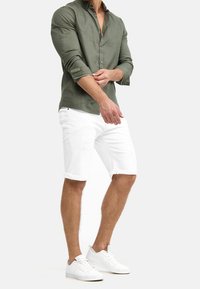 Olijfgroen overhemd met opgerolde mouwen, gecombineerd met witte denim shorts met subtiele scheuren, afgewerkt met witte sneakers.