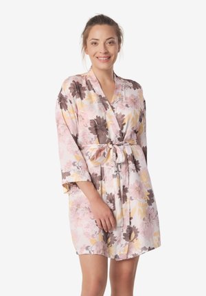 Kobieta uśmiechająca się, ubrana w jasnoróżowy szlafrok w kwiatowy wzór z przewiązanym paskiem w talii, stojąca na tle jednolitego białego tła.