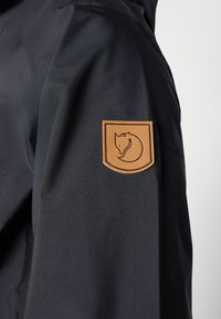 Sort jakkeærme med en brun læderlap, der har et stiliseret rævlogo broderet på overarmen mod en ensfarvet baggrund.