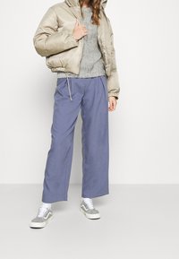Veste beige matelassée, pull en tricot gris, pantalon bleu ample et baskets grises avec lacets blancs ; texture lisse, design décontracté.