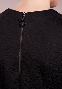 Musta teksturoitu vaate, jossa on hienovarainen kuvio, keskuselkävetoketjulla, jossa on pyöreä vetotappi, joka helpottaa pukemista.