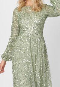 Grön paljettklänning med långa, genomskinliga ärmar, rund urringning och flödande kjol. Den texturerade tyget skapar en glittrande effekt.