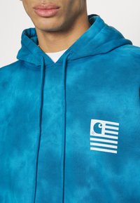 Homme portant un sweat à capuche tie-dye bleu avec un logo graphique blanc sur la poitrine et une chemise blanche en dessous.