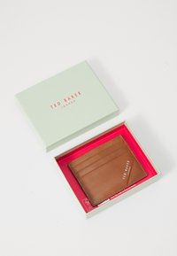 Smeđa kožna futrola za kartice s četiri utora, unutar mint zelene kutije s crvenim oblogom, s "TED BAKER LONDON" logotipom u bijeloj boji.