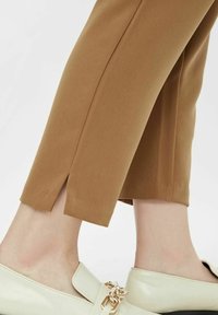 Spodnie w kolorze wielbłądzim z bocznym rozcięciem u dołu, połączone z kremowymi mokasynami zdobionymi złotym łańcuszkiem. Gładka faktura tkaniny.