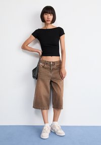 Fekete, rövidujjú crop top barna, bő szabású farmer sorttal és világos színű sportcipővel; a modell egy kis fekete kézitáskát tart.