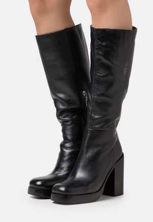 Sorte læderstøvler op til knæet med blokhæle, båret på krydsede ben, der viser lynlåsdetalje på den indvendige side af venstre støvle.