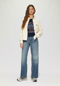 Lichtbeige corduroy jack over een donkerblauwe graphic t-shirt met bloemenpatroon, gecombineerd met lichtblauwe wijde spijkerbroek en zilveren sneakers.