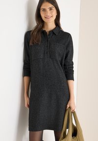 Vestido camisa negro con estampado, cuello, mangas largas y cierre de botones, combinado con un bolso verde oliva. Diseño texturizado hasta la rodilla.