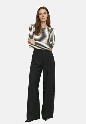Donna in piedi con le braccia incrociate che indossa un top a maniche lunghe a righe bianche e nere e pantaloni neri a vita alta e gamba larga.