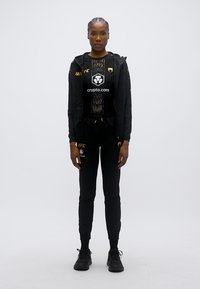 Μαύρο αθλητικό σύνολο με ζακέτα με φερμουάρ και παντελόνια με στενή εφαρμογή. Σχεδιασμένο με διακριτικά στοιχεία από δίχτυ και λογότυπα. Υφή υφάσματος, φορεμένο με αθλητικά παπούτσια.