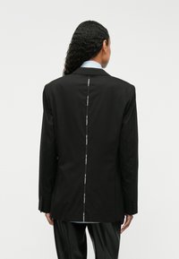 Čierny sakový blazer s klopami, jedným rozparkom vzadu a kontrastným švom a gombíkmi na manžetách, nosený cez svetlomodrú košeľu.