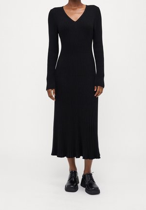 Kobieta ubrana w czarną sukienkę z dzianiny o długim rękawie z dekoltem w szpic oraz czarne platformy sznurowane, stojąca na białym tle.