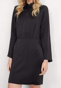 Svart knäppt klänning med långa ärmar, slät textur, figursydd bodice och utsvängd kjol. Krage med fem knappar längs framsidan.