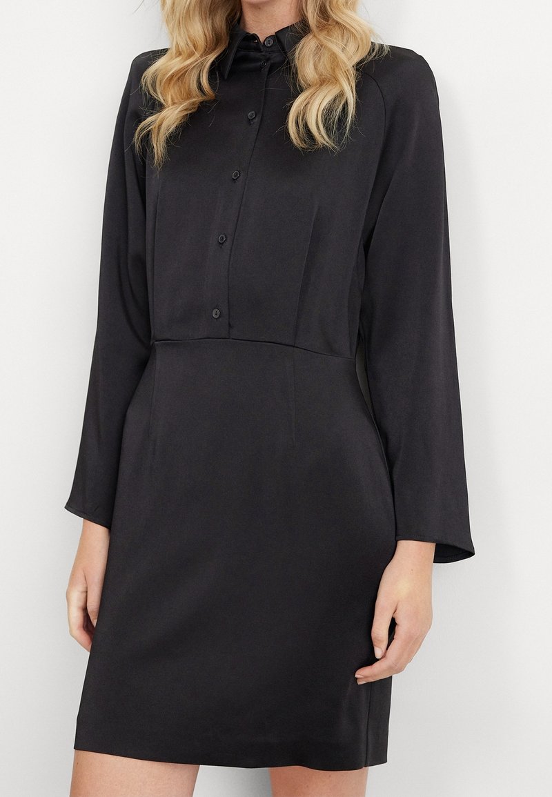 Svart knäppt klänning med långa ärmar, slät textur, figursydd bodice och utsvängd kjol. Krage med fem knappar längs framsidan.
