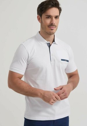 Homme aux cheveux courts et foncés portant un polo blanc à manches courtes avec un détail bleu sur la poche et le col, debout sur un fond uni.