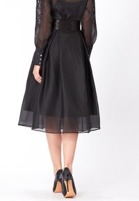 Robe noire avec des manches longues en transparent, taille ajustée et jupe évasée. Dotée de détails en boutons sur les manches et d'une texture similaire à de la satin.