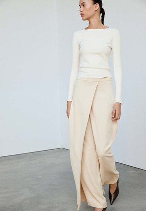 Femme aux cheveux tressés portant un haut blanc à manches longues et un pantalon large beige à nouer avec des chaussures noires à bout pointu, debout à l’intérieur.