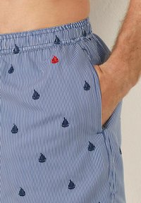 Costume de baie cu dungi albastre și albe, decorate cu mici designuri brodate de bărcuțe și un accent roșu. Talie elastică cu buzunare laterale.