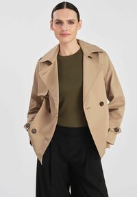 Beżowy prochowiec z dużymi klapami, podwójnym zapięciem na guziki i gładką teksturą. W połączeniu z ciemnozielonym swetrem i czarnymi spodniami.