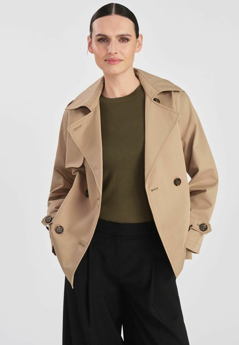 Beżowy prochowiec z dużymi klapami, podwójnym zapięciem na guziki i gładką teksturą. W połączeniu z ciemnozielonym swetrem i czarnymi spodniami.