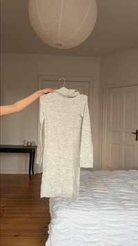 Φόρεμα με γκρι ανοιχτό χρώμα, μακρυμάνικο με ψηλό γιακά, κρεμασμένο σε κρεμάστρα που κρατιέται από το χέρι μέσα σε ένα λιτά επιπλωμένο υπνοδωμάτιο με ξύλινο πάτωμα και λευκά κλινοσκεπάσματα.