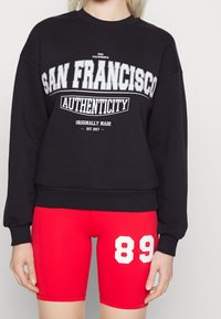 Sudadera negra con el texto blanco "AUTENTICIDAD DE SAN FRANCISCO" en la parte frontal, combinada con pantalones cortos ajustados rojos que presentan el número "89" en blanco.