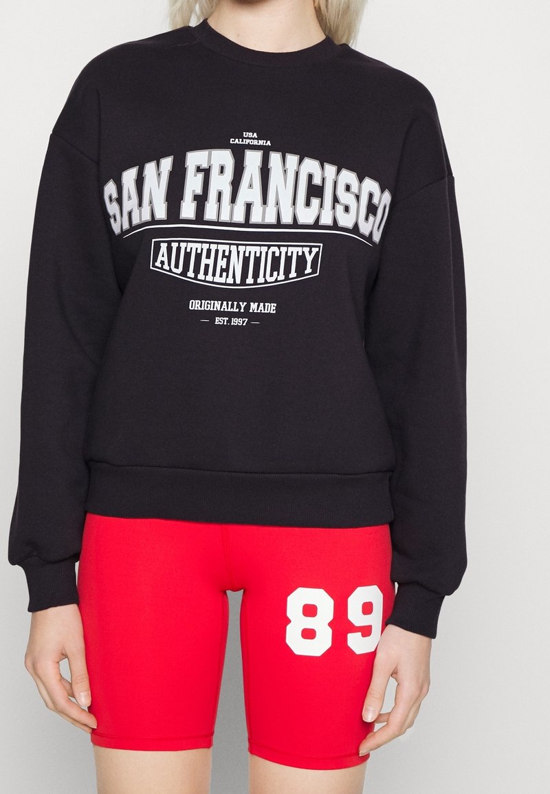 Sudadera negra con el texto blanco "AUTENTICIDAD DE SAN FRANCISCO" en la parte frontal, combinada con pantalones cortos ajustados rojos que presentan el número "89" en blanco.