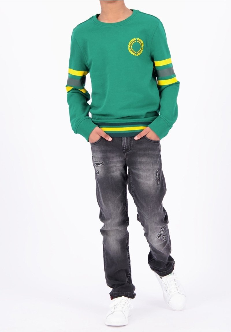 Enfant portant un sweat-shirt vert avec des rayures jaunes et un jean noir délavé, debout les mains dans les poches et des baskets blanches.