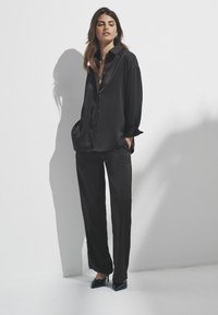 Kvinde står mod en hvid væg iført en løs sort satin skjorte, matchende vidde bukser og sorte spidse hæle, med hænderne i lommerne.