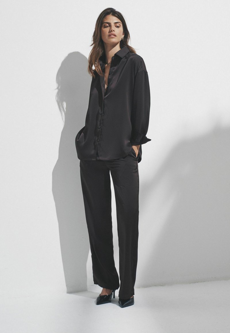 Kvinde står mod en hvid væg iført en løs sort satin skjorte, matchende vidde bukser og sorte spidse hæle, med hænderne i lommerne.