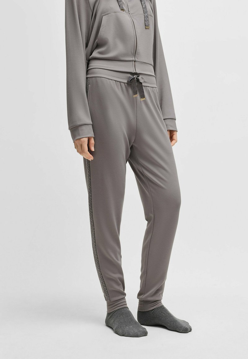 Grijze joggingbroek van zachte stof, met een koord in de taille, zijkanten met strepen en ribbelmanchetten. Draagt grijze sokken.